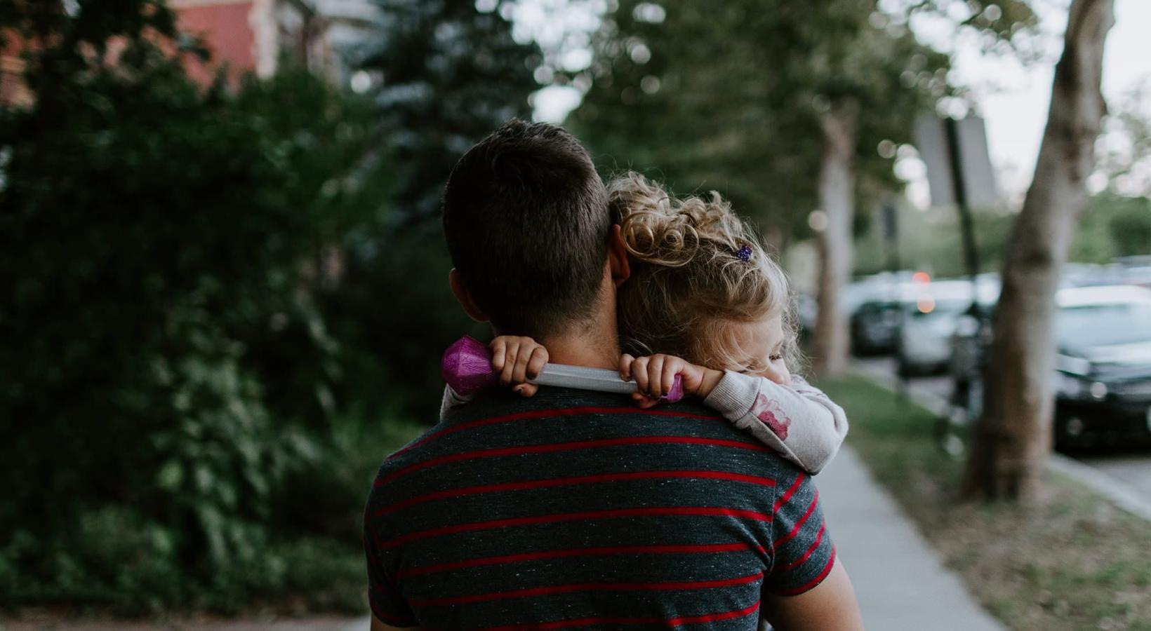 Close to the center of everything tired little girl rests her head on her father's shoulder as he carries her down the city sidewalk