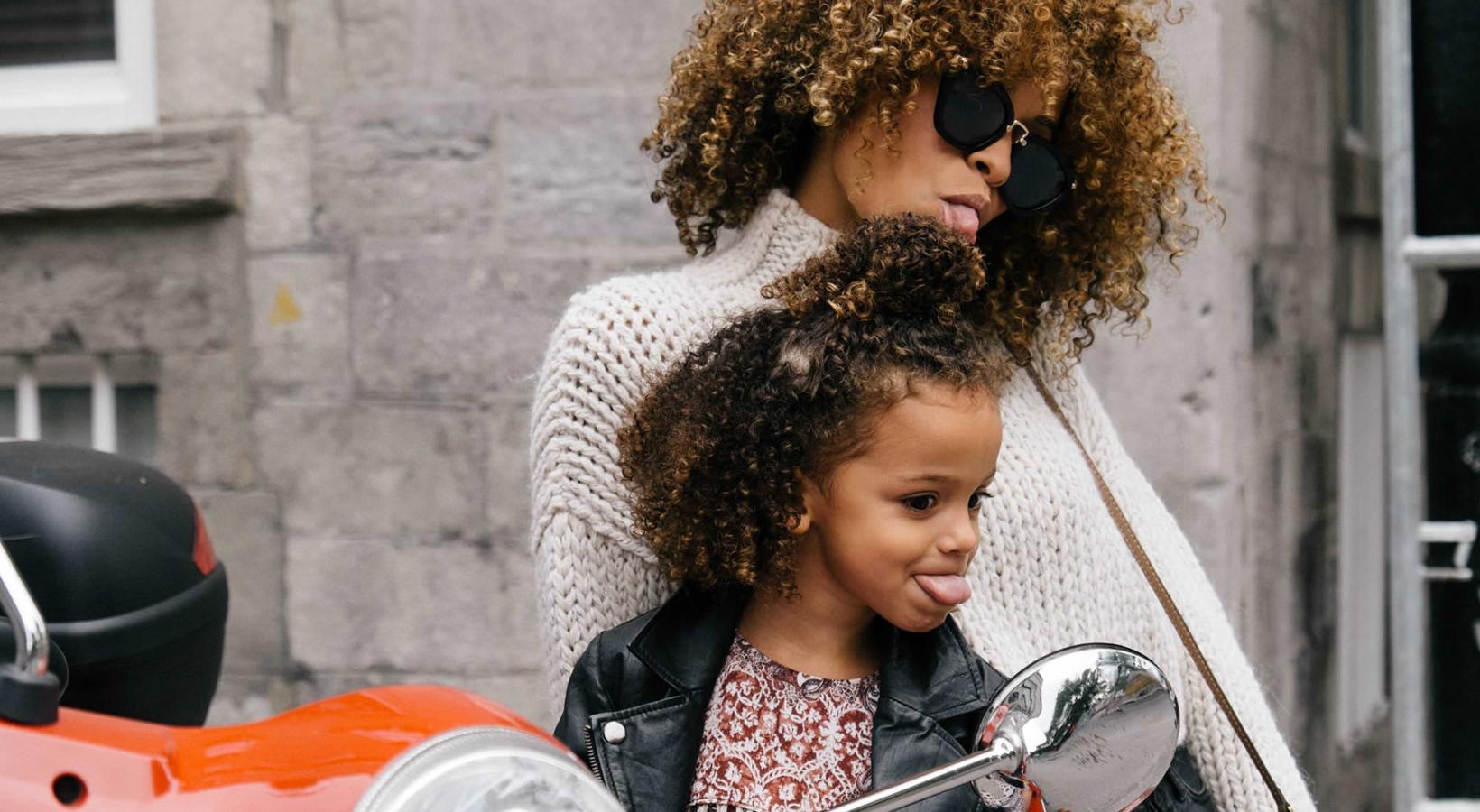 Close to the center of everything mother and daughter stick their tongues out while looking at themselves in a motorcycle mirror parked in front of a building